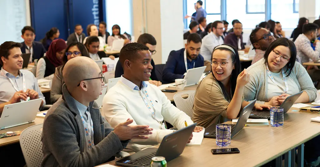 Image of a group of tech students with laptops at a long table talking in a classroom